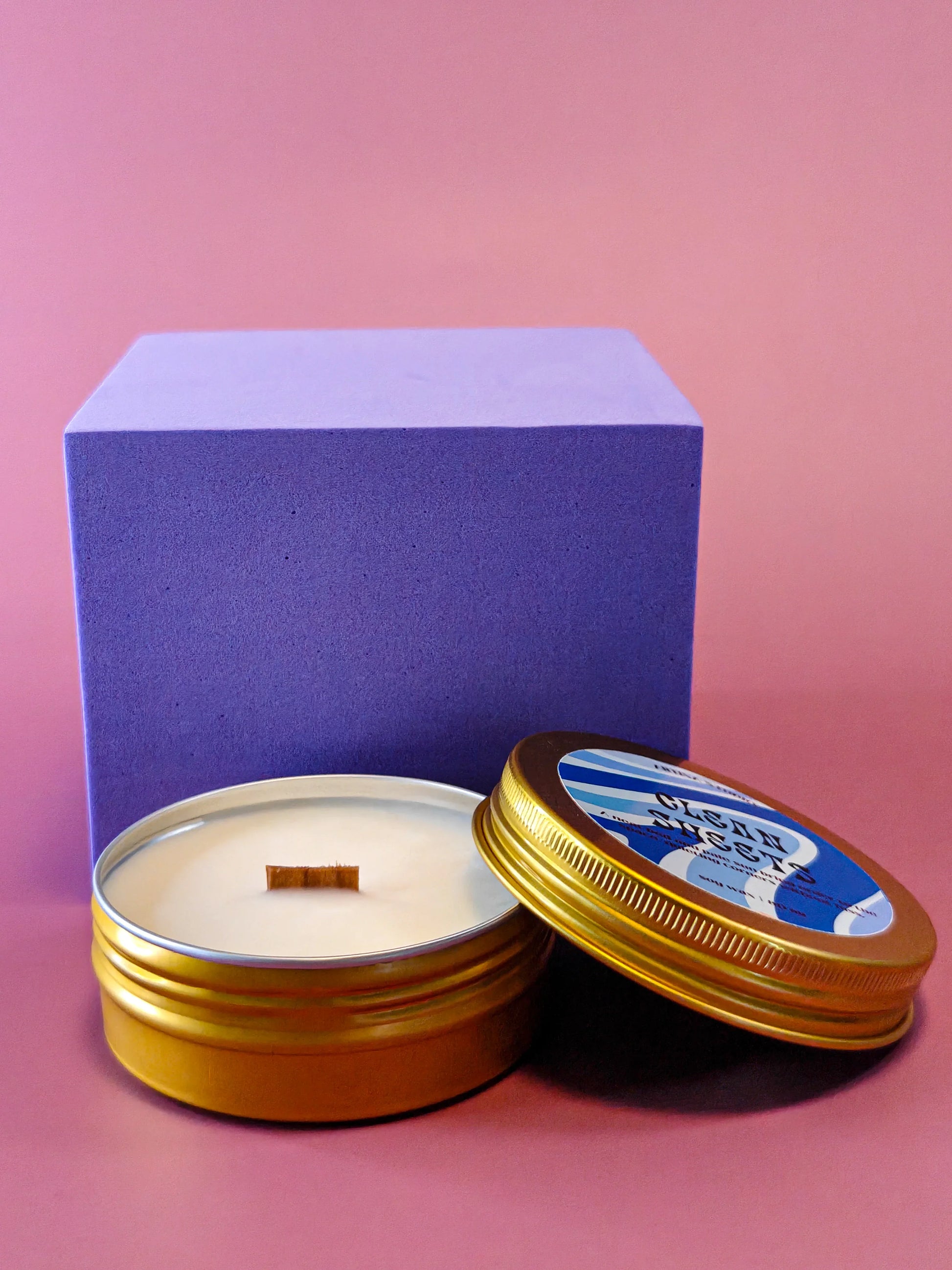 Gouden blikje met geurkaars en houten lont, paarse kubus, roze achtergrond