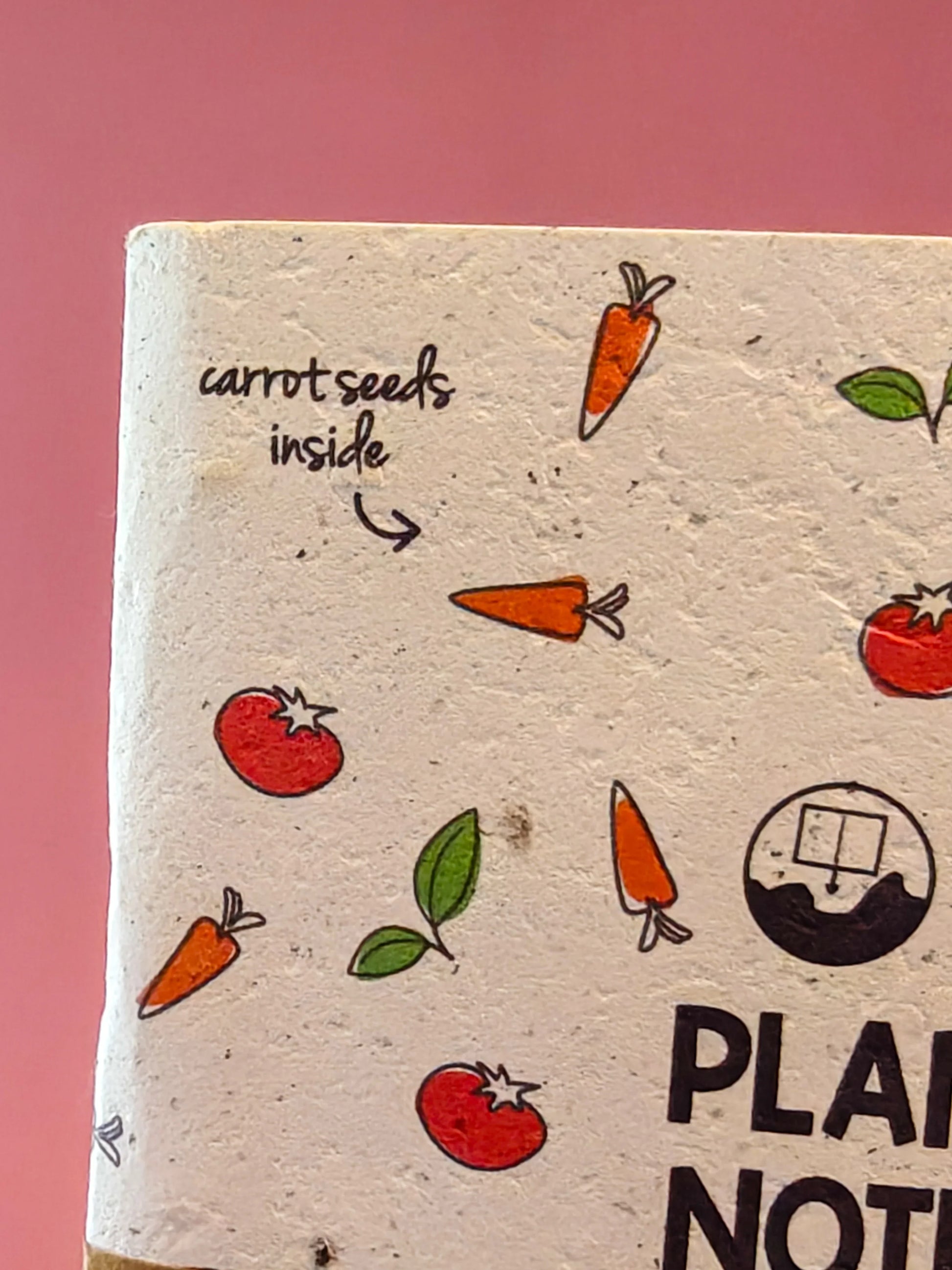 Milieuvriendelijk notitieboek van gerecycled papier met illustraties van wortels, tomaten en tekst carrot seeds inside tegen een roze achtergrond.