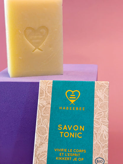 Geel blok natuurlijke zeep en verpakking 'Savon Tonic' met logo op kleurrijke achtergrond.