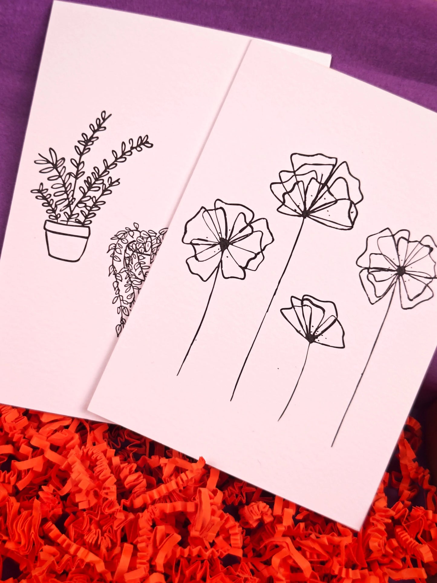Twee kaartjes met zwarte lijntekeningen van bloemen en planten, op paars en rood papier.