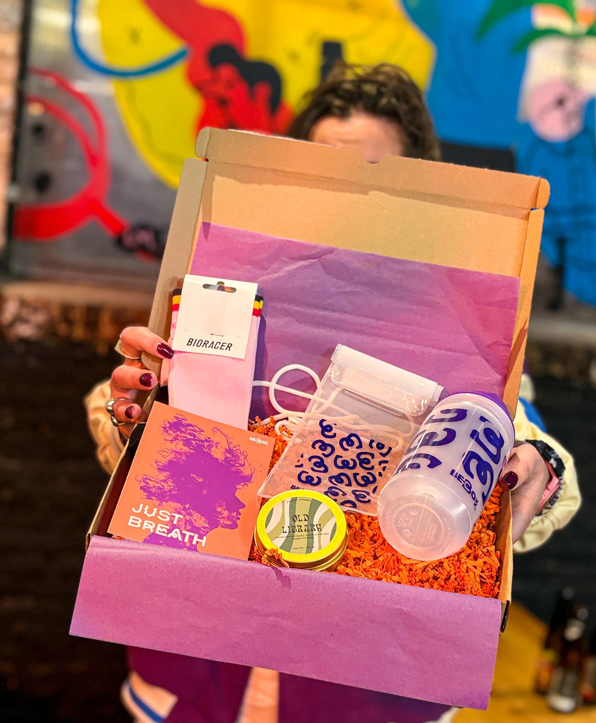 Cadeaubox met sportbidon, sokken, blikje, kaart en items op paarse vulling, tegen kleurrijke achtergrond.