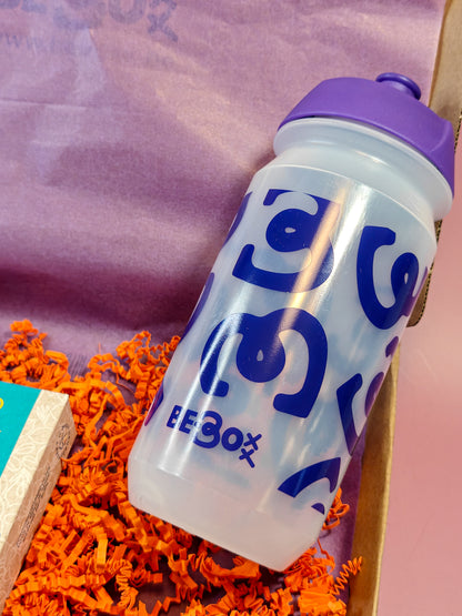 Transparante drinkfles met paarse dop en blauw patroon in geschenkbox met oranje opvulmateriaal.